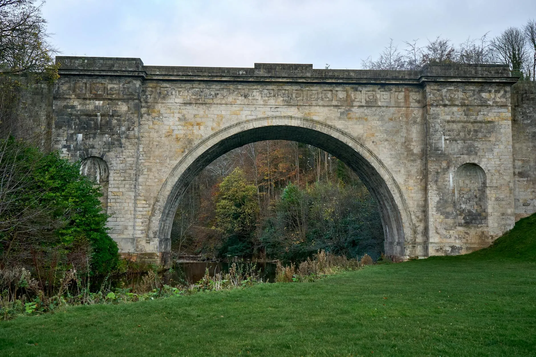 A historic stone arch bridge set in a scenic rural park with lush greenery and an overcast sky. — covered by Valley Upholstery's Stalybridge route